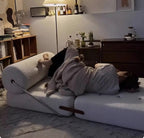 Person lying on a white couch with a blanket, surrounded by books and decor in a cozy room.