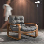 Gray cushioned armchair with wooden frame in a room with a blurred person and lamp in the background.