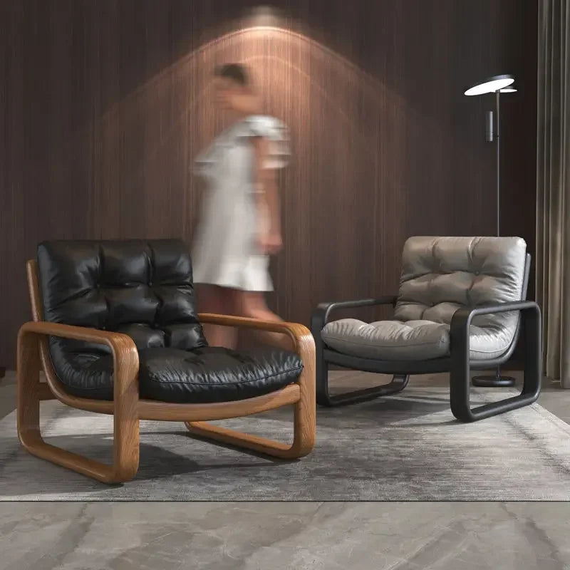 Two armchairs, one black leather and wooden frame, the other gray cushioned, in a room with a blurred person in the background.
