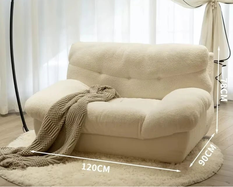 Beige armchair with a blanket draped over it, placed on a wooden floor with white curtains in the background.